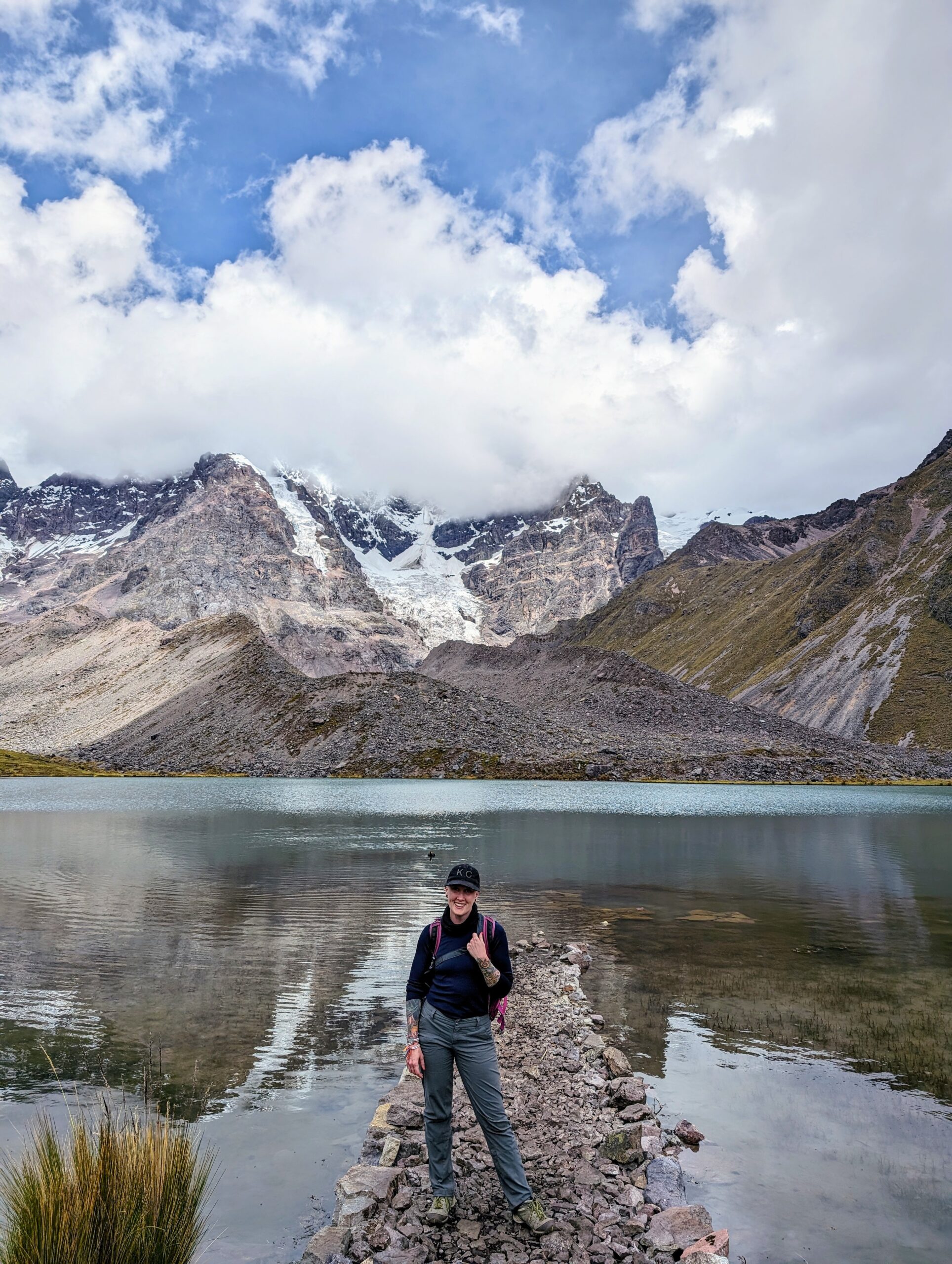Heather Physioc in Peru: A woman wearing a black cap, navy long-sleeve shirt, and gray hiking pants stands outdoors in a cloudy, snowy mountainous landscape high in the Andes near Ausangate.