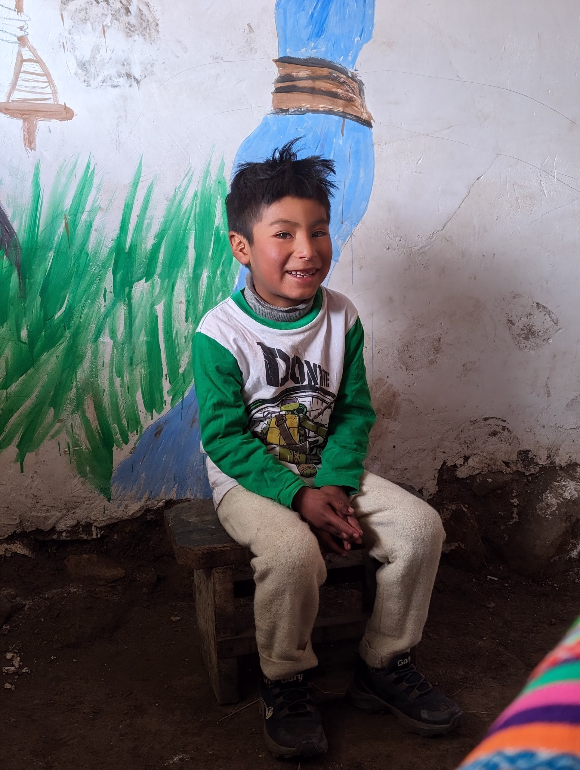 4-year-old Abelito, a Quechua child in the Andean highlands: A young Quechua boy with tousled black hair and a big smile sits on a small wooden stool inside a rustic classroom. He wears a green-and-white long-sleeve shirt featuring a cartoon turtle and fleece pants. The wall behind him is painted with childlike murals of grass and a blue figure, and the earthen floor and rough plaster add to the room’s handmade, humble charm.