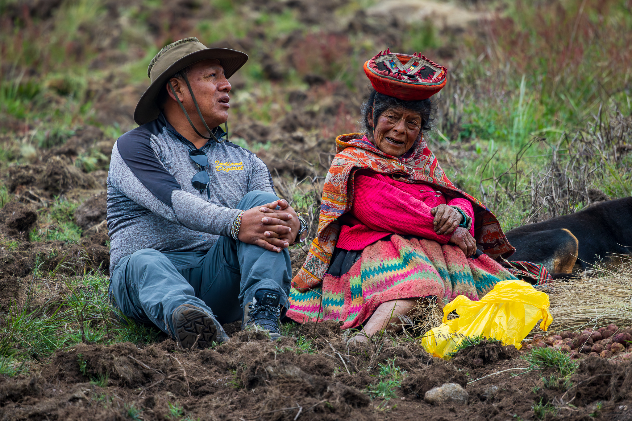A man wearing a wide-brimmed hat, hiking clothes, and boots sits on the ground beside an elderly Quechua woman dressed in a bright red sweater, patterned skirt, and traditional woven shawl. They share a quiet conversation in a highland field, surrounded by freshly turned earth, a yellow plastic bag, and scattered potatoes. A black dog rests nearby, and the soft mountain landscape fades into the background, conveying a moment of warmth and connection between travelers and locals in the Peruvian Andes.