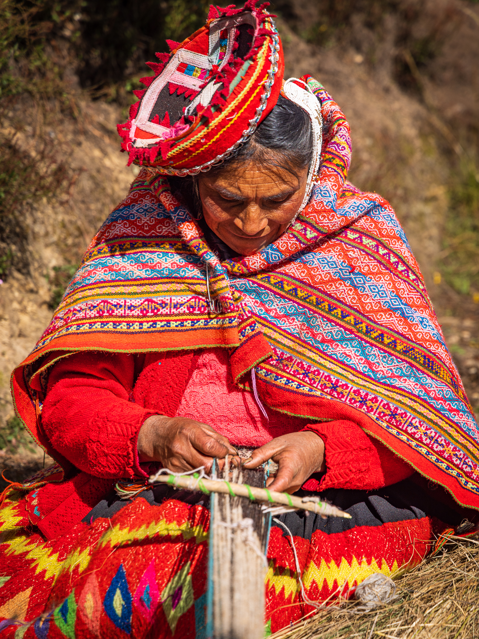 Quechua abuela hand-weaves alpaca wool using traditional methods: An elderly Quechua woman sits outdoors weaving on a traditional backstrap loom, her focus steady on the intricate threads between her hands. She wears a vivid red sweater and a handwoven shawl decorated with detailed geometric patterns in bright pink, orange, blue, and yellow. A traditional round hat adorned with ribboned edges rests on her head, tilted by the sun. The golden light highlights the textures of her clothing and the craftsmanship of her weaving—an art form passed down through generations of Andean women.