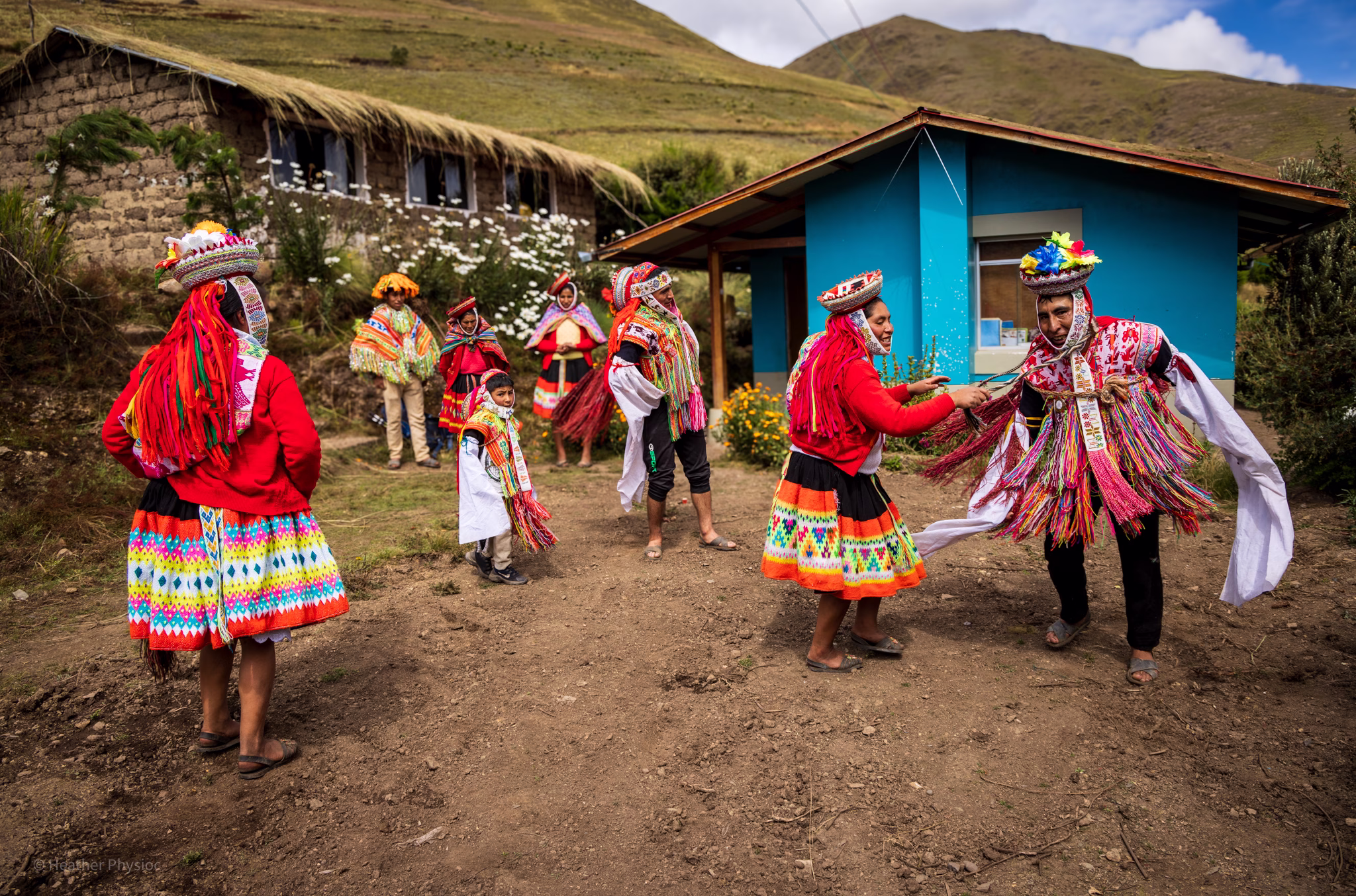 Quechua Welcome Dance: A lively outdoor scene in a rural Andean village shows men, women, and children dressed in bright traditional garments dancing and laughing near adobe houses with thatched roofs and a turquoise-painted building. The participants wear layered skirts, woven belts, and headdresses adorned with multicolored tassels and ribbons. Mountains rise in the background under a partly cloudy sky, creating a joyful, celebratory atmosphere.