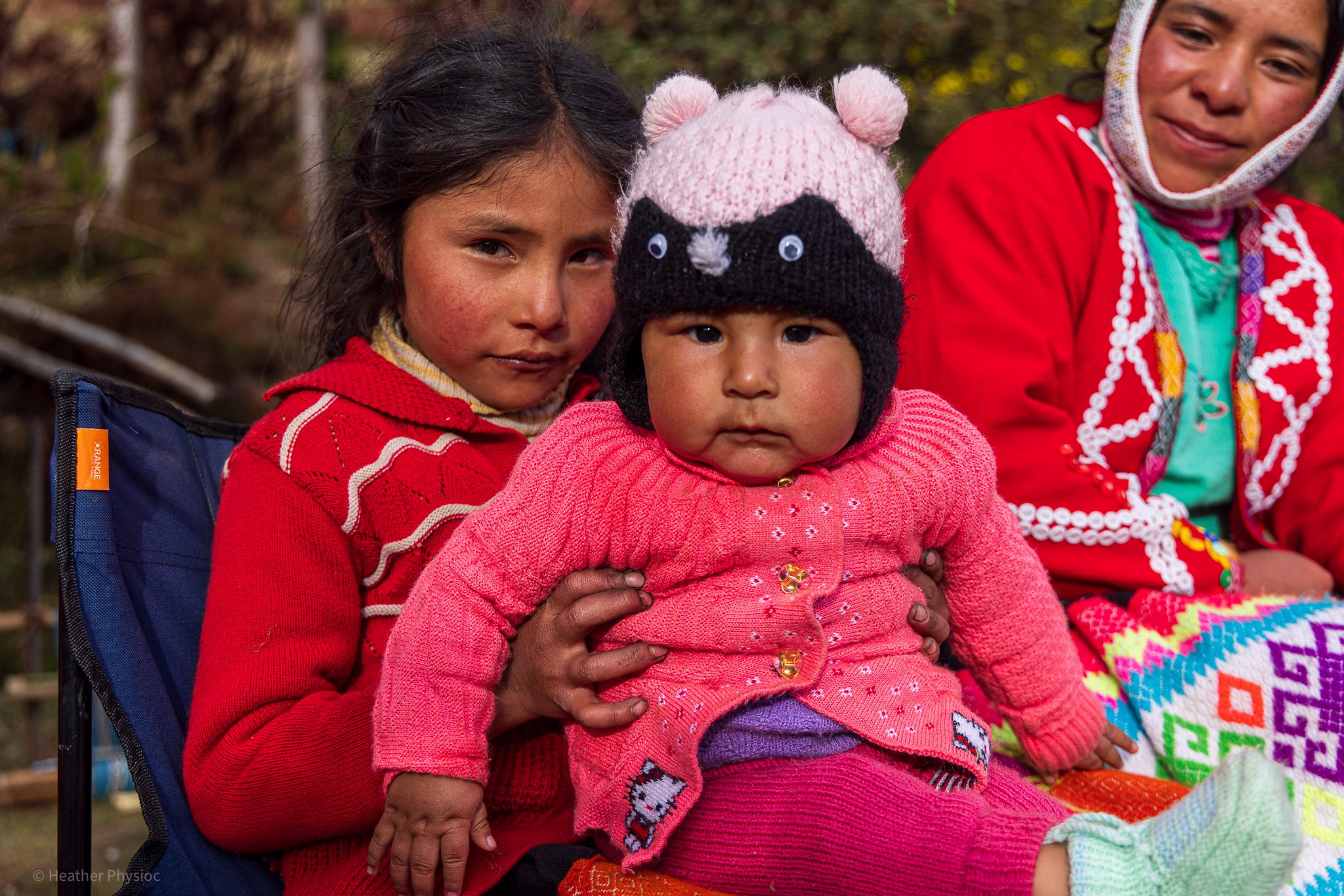 A young Quechua girl wearing a red sweater holds a bundled baby dressed in pink knit clothing and a hat with animal ears and button eyes. Both sit outside in soft natural light, their cheeks flushed from the mountain air. An older woman in the background, also in traditional colorful Andean clothing, watches with a gentle smile. The image captures the warmth and shared caregiving that are central to family life in Quechua communities.