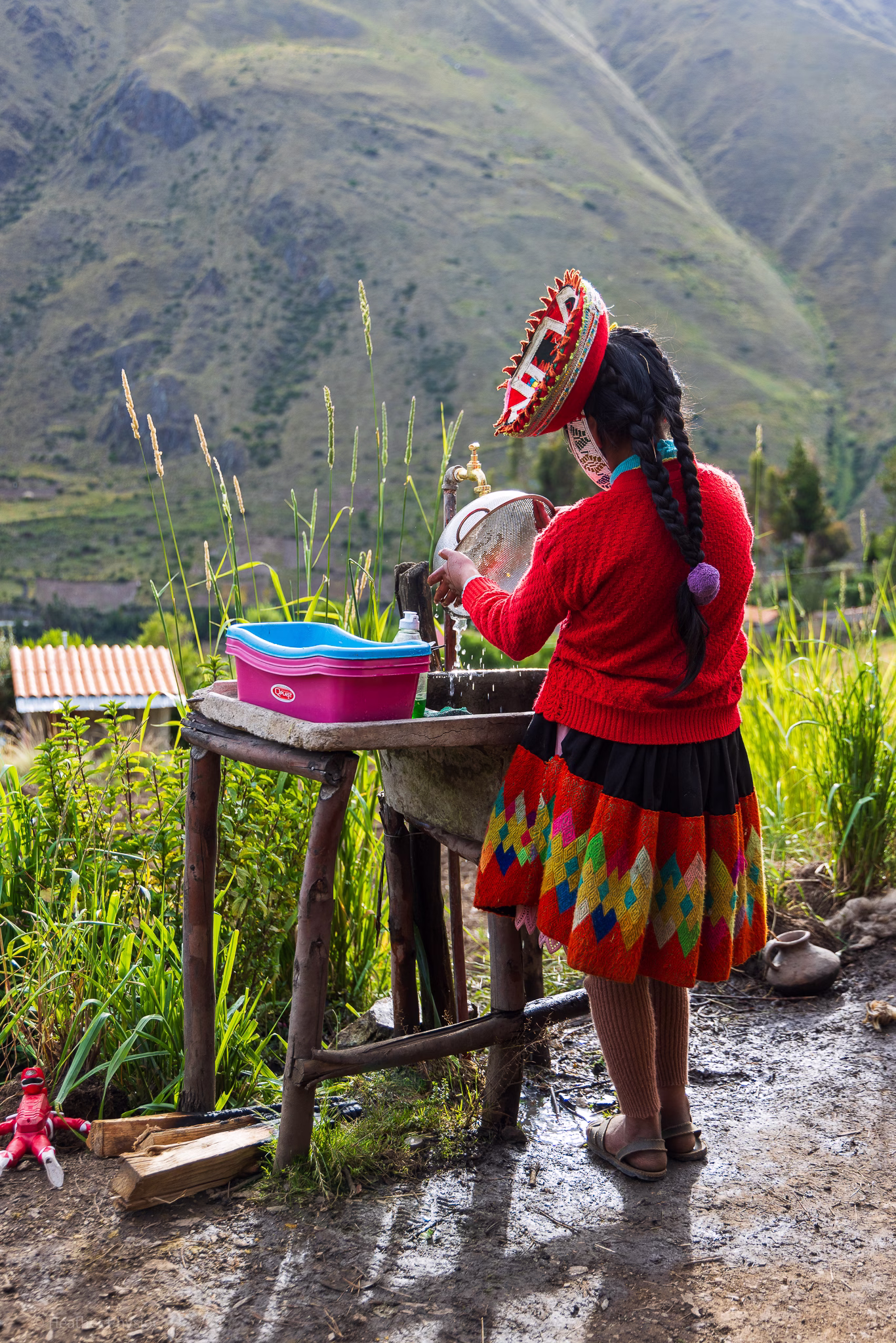 Young Quechua woman washes dishes in the shadows of the Andes: A young Quechua girl, wearing a traditional red sweater, black skirt with bright geometric patterns, and a round embroidered hat, stands at an outdoor sink washing dishes. Water trickles from a brass faucet as she cleans a metal bowl beside a pink plastic basin. Her long braids fall down her back as late afternoon light illuminates the green mountain slopes surrounding her rural home near Patacancha in the Peruvian Andes.