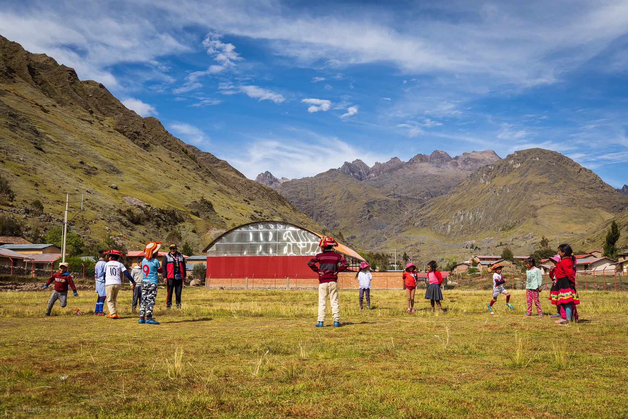 A group of Quechua schoolchildren plays a supervised outdoor game on a grassy field surrounded by towering green mountains in the Peruvian Andes. The children wear a mix of traditional Andean clothing—bright red sweaters, woven skirts, and hats—and modern sportswear with soccer jerseys and sneakers. A teacher stands nearby overseeing the activity, while a red-roofed school building and gymnasium glint in the sunlight under a vivid blue sky,.