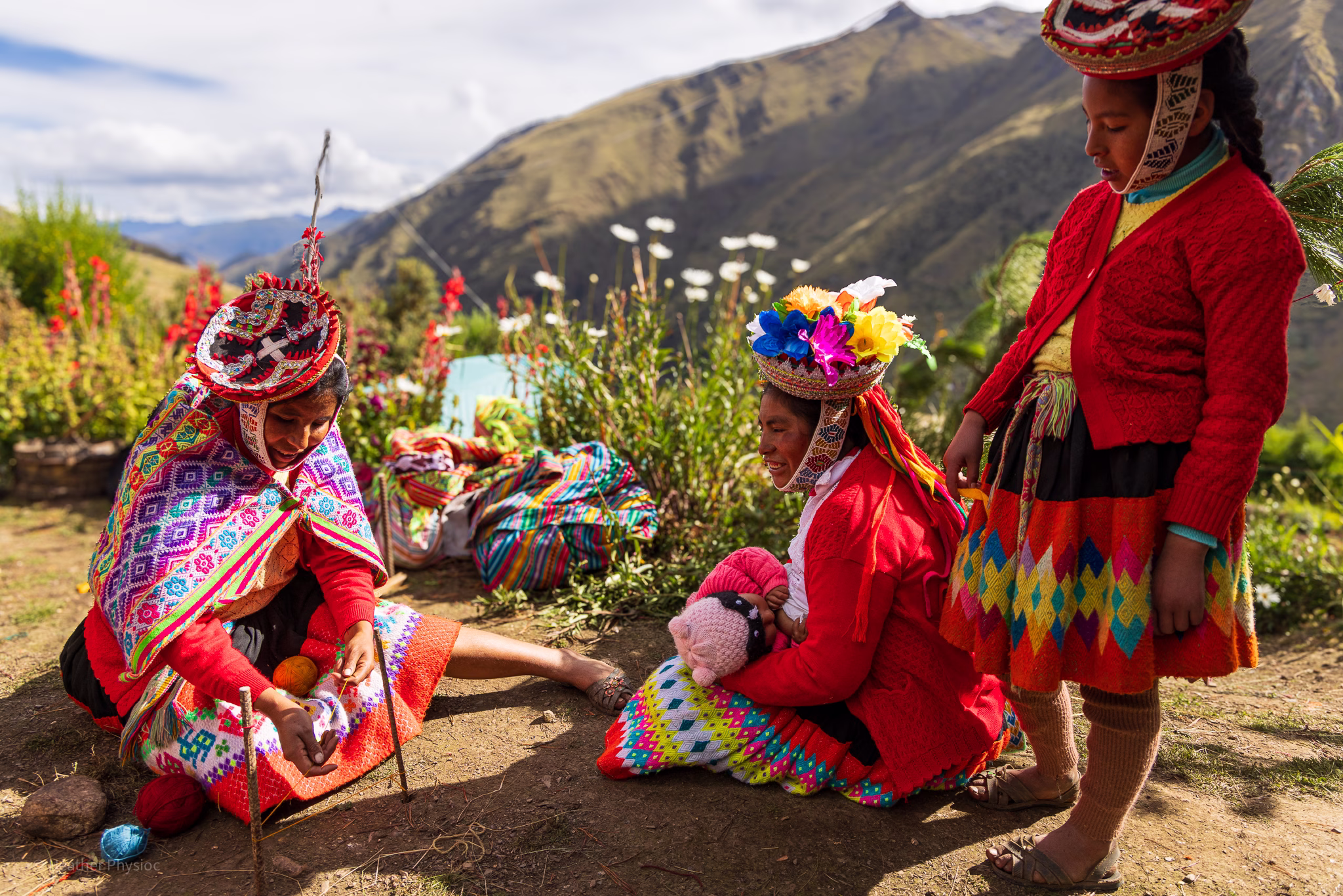 Three Quechua women, one a young teenager and two adult women weaving, with a young baby breastfeeding. All are dressed in brightly dyed wools of red, pink, yellow and purple, from their alpaca weaving. Three Quechua women and girls sit and stand outdoors in the Andean highlands, surrounded by blooming flowers and mountain views. They wear traditional handwoven clothing—bright red sweaters, patterned skirts, and elaborately embroidered hats adorned with ribbons and flowers. One woman sits cross-legged on the ground spinning colorful wool, while another cradles a baby dressed in pink. The warm sunlight highlights the vivid textiles and the women’s deep connection to their heritage and landscape.