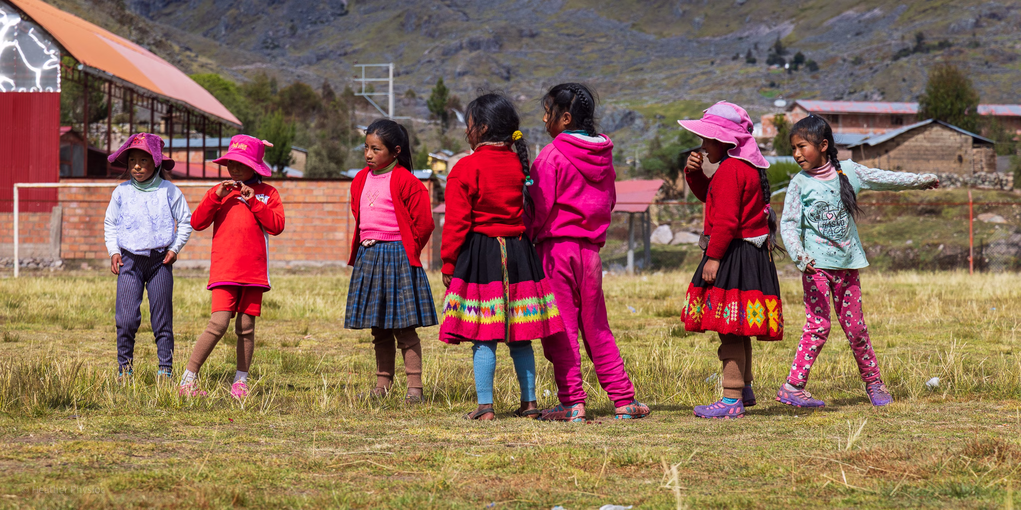 A group of eight Quechua schoolgirls stands together on a grassy field in the Andean highlands, playing an outdoor game under the mountain sun. They wear bright clothing — red sweaters, colorful skirts, pink hats, leggings, and sneakers — reflecting both traditional Andean patterns and modern styles. A few look at one another mid-conversation, while others glance toward the camera. Behind them are rustic brick school buildings and a corrugated-roof structure, with rocky mountain slopes rising in the background.