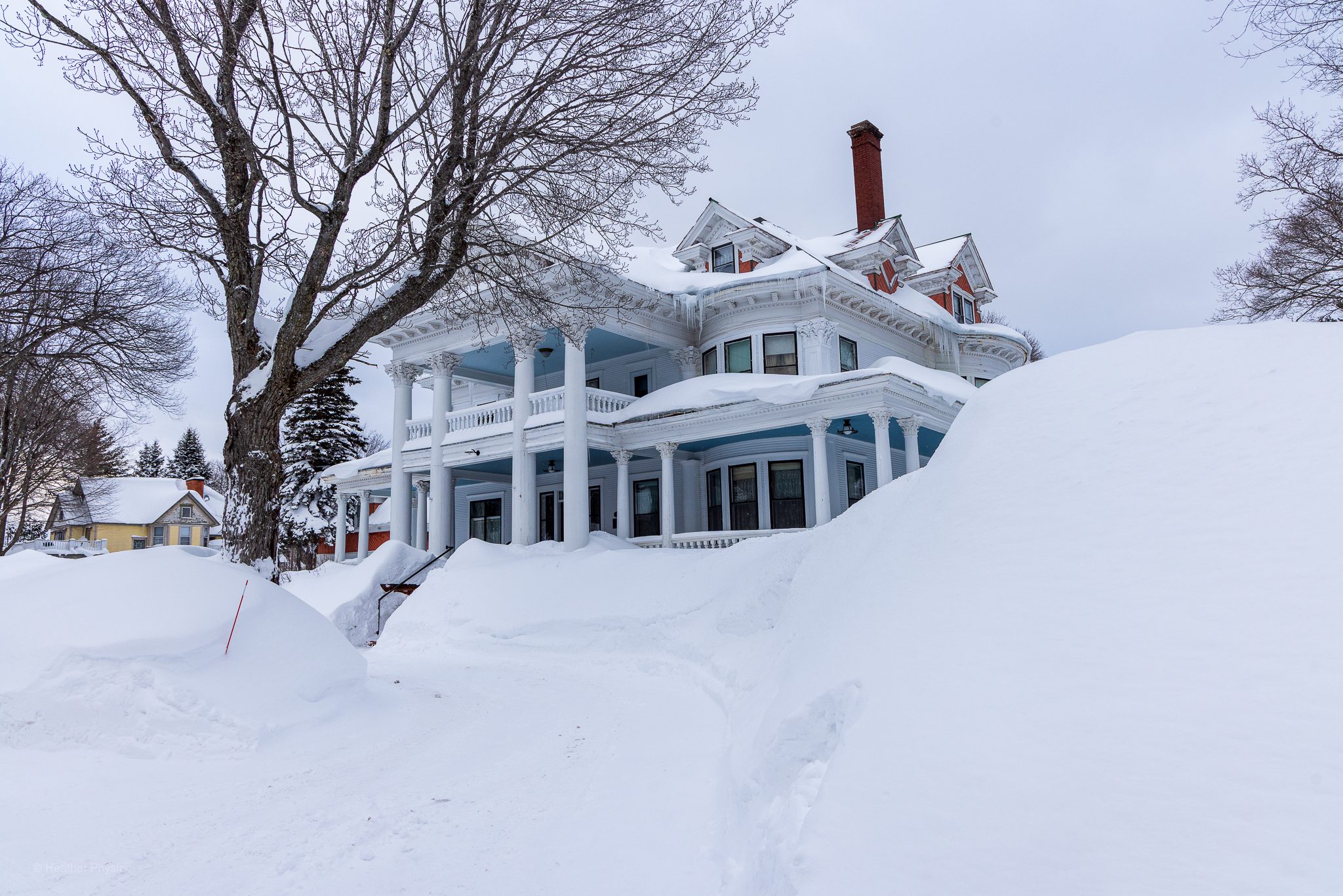 The historic Laurium Manor Inn stands amid towering snowbanks during winter in Michigan’s Upper Peninsula. The large white mansion features grand columns, wraparound porches, ornate Victorian-era architectural details, and a tall brick chimney, with heavy snow piled high around the property beneath an overcast sky.