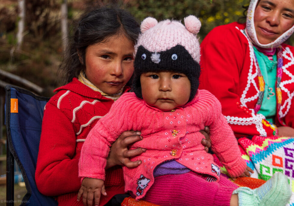 quechua-language-revitalization-story-take-1-09-1 A young Quechua girl in a red sweater sits on a blue chair outdoors, holding a bundled baby dressed in a pink knit sweater and a black-and-white animal-ear hat. The baby looks directly at the camera with a serious expression, while an older woman in colorful traditional clothing and a white woven head covering smiles softly beside them. The scene is warmly lit, showing textures of wool, embroidery, and rosy cheeks against a soft, natural background.